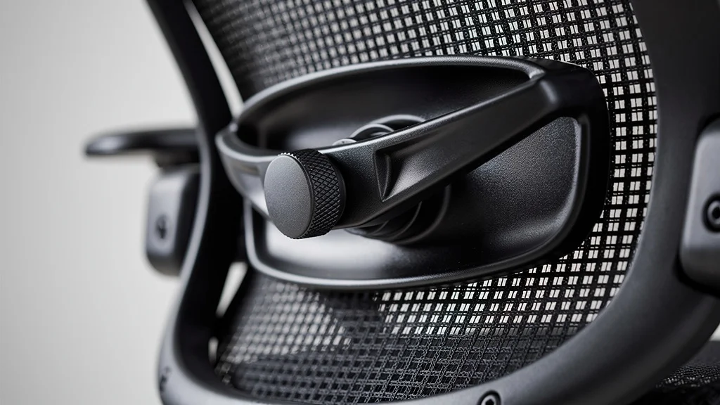 Extreme close-up macro photograph of a black mesh office chair backrest showing the adjustable lumbar support mechanism, knob and pivoting plastic — engineering detail of what separates real ergonomic chairs from fake ones